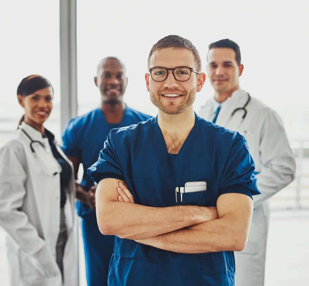 Males nurse in foreground with doctors in background smiling.